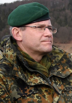 Nahaufnahme eines Mannes mittleren Alters mit grünem Barett und Flecktarn-Uniform, Brille tragend, nach rechts blickend.