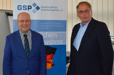 Zwei lächelnde Männer in Anzügen stehen vor einem Banner der "Gesellschaft für Sicherheitspolitik e.V.".
