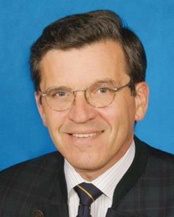 Porträt eines lächelnden Mannes mit Brille, dunklem Haar, weißem Hemd, gestreifter Krawatte und dunklem Sakko vor blauem Hintergrund.