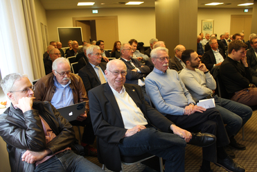 Eine Gruppe von überwiegend Männern sitzt als Publikum in einem Konferenzraum. Ein älterer Herr lächelt in die Kamera.