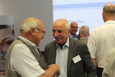 Zwei lächelnde Senioren, Jürgen Höche unter ihnen, unterhalten sich auf einer Veranstaltung mit weiteren Gästen.