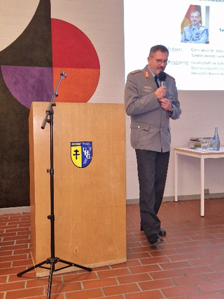 Ein Militäroffizier in Uniform spricht in ein Mikrofon an einem Rednerpult mit einer Präsentationsleinwand dahinter.