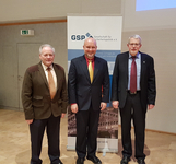 Drei Männer in Anzügen stehen bei einer GSP-Veranstaltung vor einem Banner und einem Podium mit Bundesadler.