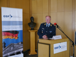 Dr. Heiner Möllers, ein Offizier, lächelt am GSP-Rednerpult. Ein Banner mit deutscher Flagge und Reichstag ist im Hintergrund.