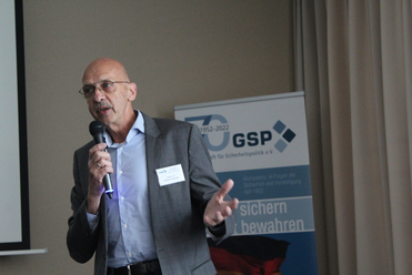 Brig. Gen. a.D. Reiner Schwalb spricht auf einer GSP-Veranstaltung ins Mikrofon. GSP-Banner mit Deutschlandflagge hinten.