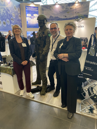 Drei Personen stehen an einem Verteidigungstechnik-Messestand neben einer Militärmannequin, mit Displays im Hintergrund.