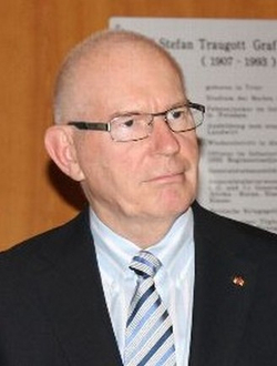 Kahlköpfiger Mann mit Brille in Anzug und gestreifter Krawatte, blickt leicht nach rechts. Ein Schild im Hintergrund.