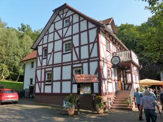 Ein traditionelles Fachwerkhaus namens Ziegelhütte mit rotem Auto und Besuchern, umgeben von Bäumen unter blauem Himmel.
