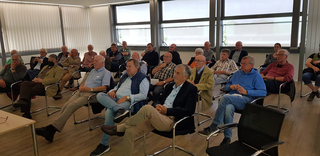 Eine Gruppe meist älterer Männer sitzt in Reihen in einem gut beleuchteten Raum und nimmt an einer Besprechung teil.