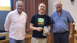 Drei lächelnde Männer, einer in einem "Taiwan is my country"-Shirt zeigt auf das Buch "Die wichtigste Insel der Welt".