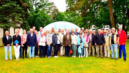 Große Gruppe von Erwachsenen und Senioren posiert lächelnd auf einer grünen Wiese vor einem hellblauen, kuppelartigen Gebäude.