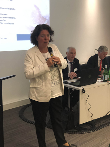 Eine Frau spricht mit Mikrofon während einer Präsentation vor einem Bildschirm und sitzenden Teilnehmern.