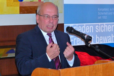 Ein kahlköpfiger Mann mit Brille im dunklen Anzug spricht an einem Holzpult und gestikuliert. Mikrofon und blaues Banner im Hintergrund.