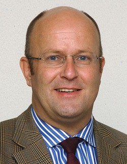 Porträt eines lächelnden Mannes mittleren Alters mit Glatze und Brille, im karierten Sakko, gestreiften Hemd und Krawatte.