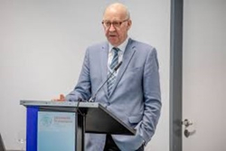 Älterer Mann in Anzug und Brille spricht an einem Rednerpult auf einer Konferenz.
