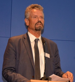 Mann mit grauem Haar und Bart im Anzug und Krawatte spricht an einem Rednerpult vor blauem Hintergrund.