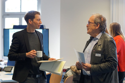 Zwei Männer, der jüngere mit Tasse und Papieren, der ältere in Lederjacke, unterhalten sich bei einer Veranstaltung.