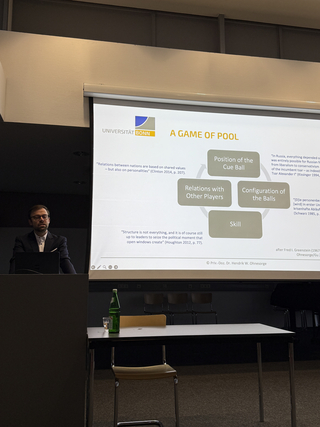 Ein Mann präsentiert eine Folie der Universität Bonn mit dem Titel "A Game of Pool" und einem Diagramm zu Politikfaktoren.