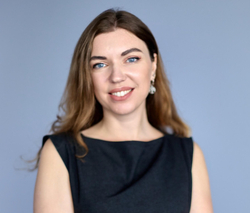 Eine lächelnde Frau mit langen braunen Haaren, blauen Augen und schwarzem Oberteil vor hellblauem Hintergrund.