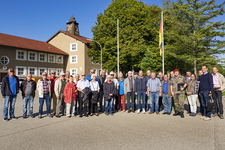 Eine große Gruppe verschiedener Personen, darunter ein Soldat, posiert draußen vor einem Gebäude mit Uhrturm und deutscher Flagge.
