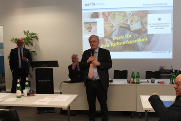 Mann hält Vortrag bei Konferenz, Projektion zeigt Euroscheine, Danksagung und Neujahrswunsch des Finanzministers.