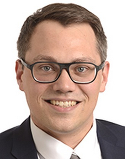 Porträt eines lächelnden Mannes mit Brille, kurzem dunklem Haar, Anzug und Krawatte, vor weißem Hintergrund.