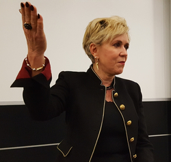 Blonde Frau in schwarzer Jacke mit Goldbesatz hebt die Hand, als würde sie einen Eid leisten oder schwören.