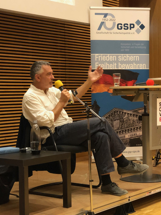 Ein Mann spricht bei einer GSP-Veranstaltung in ein gelbes Mikrofon, hinter ihm ein Banner für Frieden und Freiheit.