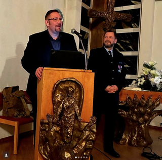 Zwei Männer in einem religiösen Raum. Einer spricht am Rednerpult, der andere im Uniform daneben stehend.
