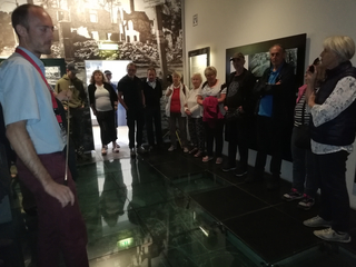 Ein Führer spricht eine Gruppe von Menschen in einem Museum mit Glasboden und großen Schwarz-Weiß-Fotos zerstörter Gebäude an.