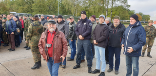 Gruppe von Personen, einige in Tarnkleidung, andere in Zivil, stehen im Freien; ein lächelnder Mann mit Kamera im Vordergrund.