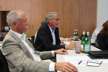 Zwei Männer an einem Konferenztisch: einer im karierten Sakko, ein anderer im dunklen Anzug, mit Papieren und Wasserflaschen.