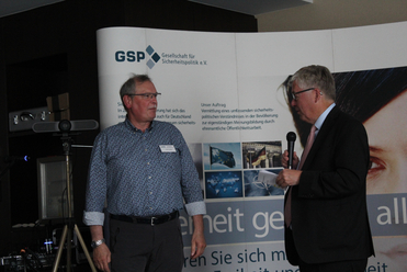 Zwei Männer, einer mit Mikrofon, stehen vor einem Banner der Gesellschaft für Sicherheitspolitik (GSP).