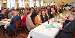 Zahlreiche Menschen bei einem festlichen Treffen in einem Saal, viele sitzend an dekorierten Tischen, einige in Uniform.