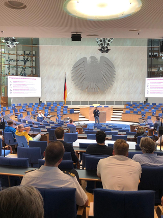 Redner in einem Hörsaal mit Bundesadler an der Wand, Präsentation zu Erfolgsfaktoren der Bundeswehrverwaltung.