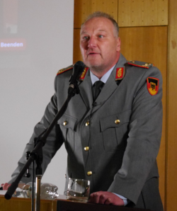 Ein Mann in grauer deutscher Militäruniform mit Deutschlandflaggen-Aufnäher spricht an einem Rednerpult mit Mikrofon.