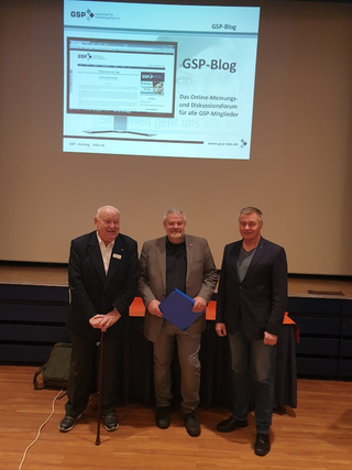 Drei Männer, darunter ein älterer Mann mit Gehstock und ein Mann mit blauer Mappe, posieren vor einer GSP-Blog Präsentation.