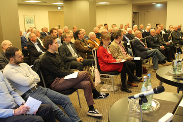 Gemischtes Publikum, hauptsächlich Männer, sitzen in einem hellen Konferenzraum und lauschen einer Präsentation. Getränke und Mikrofon.