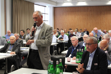 Mann mit Schnurrbart spricht ins Mikrofon auf einer Konferenz vor sitzenden Teilnehmern.