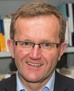 Porträt eines lächelnden Mannes mit Brille, blauem Hemd und dunklem Sakko, Bücher im Hintergrund unscharf.