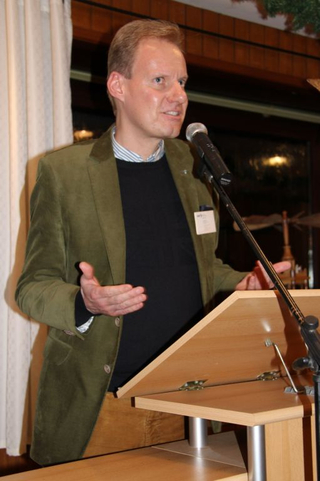 Mann in grünem Sakko hält Vortrag an einem Rednerpult mit Mikrofon, gestikuliert während der Präsentation.