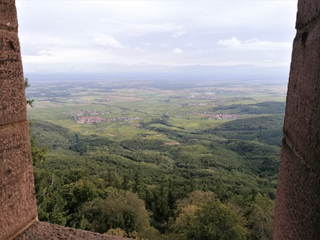 Panoramablick aus einem Burgfenster auf ein weites Tal mit Wäldern, Weinbergen, Dörfern und Feldern unter bewölktem Himmel.