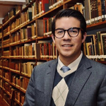 Ein lächelnder Mann mit Brille in einem Anzug mit Argyle-Weste und Krawatte steht vor Bücherregalen in einer Bibliothek.