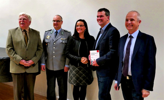 Fünf lächelnde Personen, darunter ein Soldat, in einer Reihe. Ein Mann hält ein Buch: 'Sicherheitspolitik Verstehen'.
