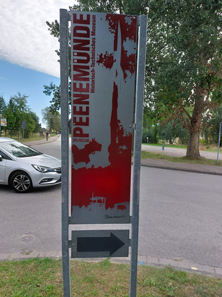 Schild: Historisch-Technisches Museum Peenemünde, mit Raketensilhouette und Pfeil nach rechts, an einer Straße.