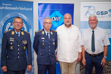 Vier Männer, davon zwei in Uniform und zwei in Zivil, stehen vor Bannern verschiedener Organisationen.