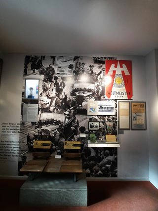 Museumsausstellung zur Fußball-WM 1954 'Wunder von Bern' mit Team-Wandbild, Trophäe, Dokumenten und alten Stadionbänken.