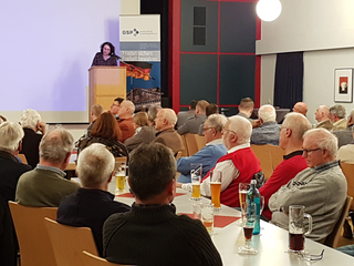 Eine Frau spricht am Rednerpult vor einem Publikum, meist ältere Männer, bei einer GSP-Veranstaltung mit Getränken auf den Tischen.