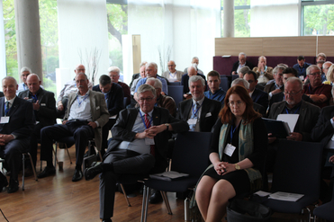 Eine Gruppe von größtenteils älteren Männern und einer Frau sitzt in einem Konferenzraum und lauscht einer Präsentation.