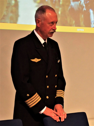 Ein älterer Mann mit Schnurrbart in dunkler Uniform mit goldener Pilotenflügel-Anstecknadel und Rangstreifen, Hände auf Stuhllehne.